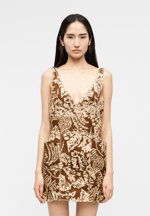 Mladá žena s dlhými tmavými vlasmi, oblečená v hnedých bezrukávových šatách s béžovými abstraktnými ručne písanými textovými vzormi a hlbokým V-výstrihom.