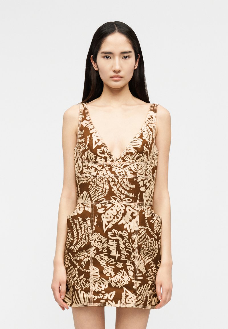 Νεαρή γυναίκα με μακριά σκουρόχρωμα μαλλιά, που φοράει ένα αμάνικο καφέ φόρεμα με μπεζ αφηρημένα γραφικά μοτίβα σαν χειρόγραφο κείμενο και βαθύ V-λαιμόκοψη.