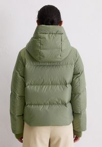 Zielona pikowana kurtka puchowa z kapturem, z przeszyciami i gładką teksturą. Nosi ją osoba w beżowych spodniach ze sztruksu, widziana od tyłu.