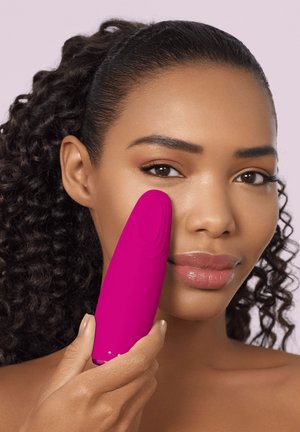 Kvinde med krøllet hår påfører en lyserød ansigtsrensningsenhed på sin kind mod en lyserød baggrund.