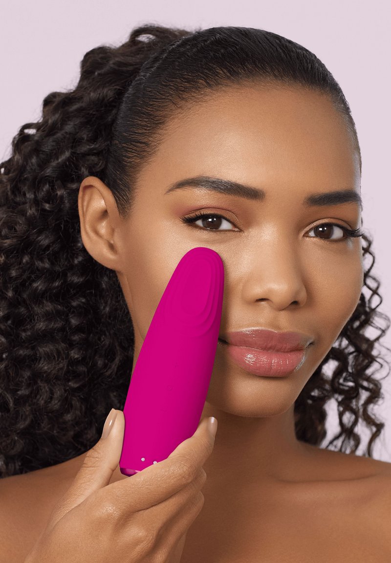 Woman with curly hair applies a pink facial cleansing device to her cheek against a pale pink background.