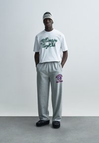 Λευκό T-shirt με πράσινο λογότυπο, γκρι φόρμα με μοβ λεπτομέρεια λογοτύπου και μαύρα παπούτσια slip-on. Απλός, καθημερινός σχεδιασμός streetwear.