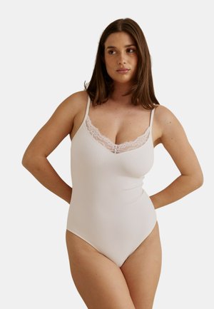 Femme portant un body camisole côtelé rose clair avec bordure en dentelle, debout les mains sur les hanches, sur un fond clair uni.