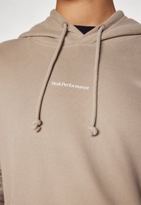 Beige hoodie met trekkoord, voorzien van een voorzijde logo "PeakPerformance" in het wit. Zachte textuur en klassiek ontwerp met een buidelzak.