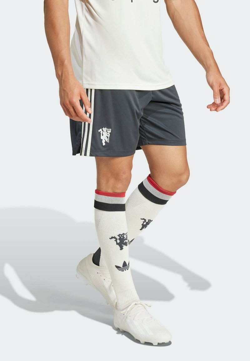 Homme portant des chaussures de sport blanches, de longues chaussettes blanches avec des rayures noires et rouges et des logos, un short de sport foncé avec des rayures blanches et un emblème.