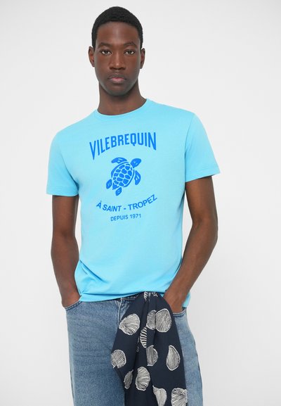 Svjetloplava pamučna majica s plavim grafikom kornjače i natpisom. Ima kratke rukave i okrugli izrez. Uparena s uzorkovanom tamnoplavom maramom.