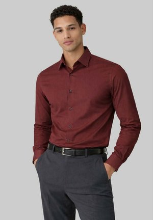 Homme portant une chemise bordeaux à manches longues, ajustée et boutonnée, rentrée dans un pantalon gris foncé avec une ceinture noire, les mains dans les poches.