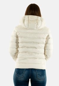 Chaqueta de plumas blanca con capucha, con costuras horizontales, textura suave y un diseño ajustado. Se lleva con jeans azules.