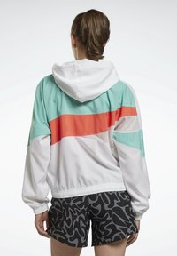 Giacca colorata con strisce orizzontali bianche, turchesi e corallo, dotata di cappuccio e dettagli in rete, abbinata a pantaloni corti neri con motivo.
