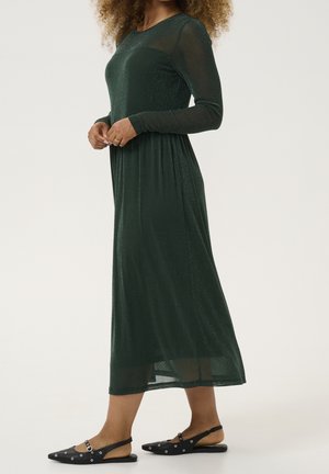 Kobieta ubrana w zieloną, długorękawową sukienkę midi z połyskiem, w połączeniu z czarnymi, spiczastymi balerinkami na pasku z srebrnymi oczkami.