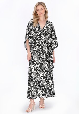 Kvinde stående iført en lang kjole med sort og hvidt blomstermønster, brede ærmer og beige remsandaler med blokhæl.