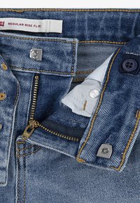 Jeans de mezclilla en azul claro con tiro regular y pierna acampanada. Presenta costuras doradas, un zipper plateado y un cierre con botón.