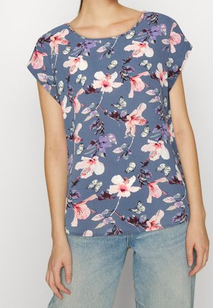 Kvinde iført en blå kortærmet top med lyserøde og lilla blomster- og sommerfuglemønstre kombineret med lyseblå jeans.