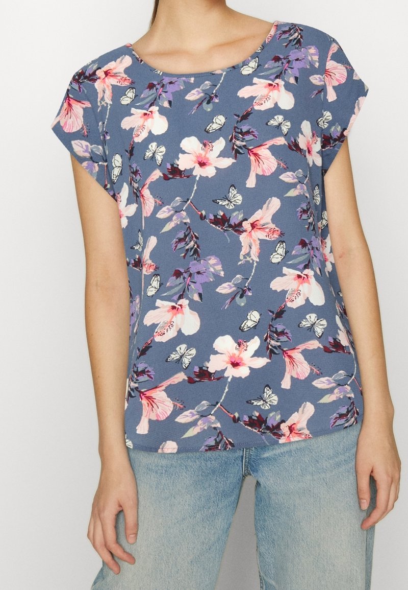 Femme portant un haut bleu à manches courtes avec des motifs floraux et papillons roses et violets, associé à un jean bleu clair.