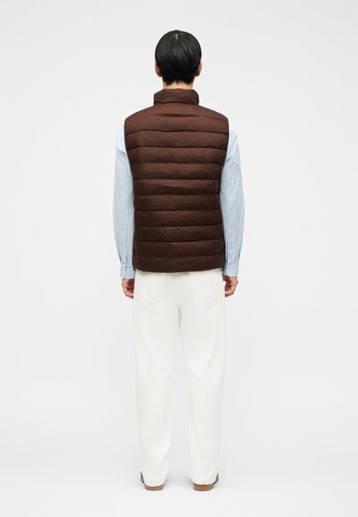 Άνδρας που φοράει καφέ στρωμένο γιλέκο, μπλε και λευκό ριγέ πουκάμισο και λευκό παντελόνι, στέκεται κοιτώντας προς ένα απλό λευκό φόντο.