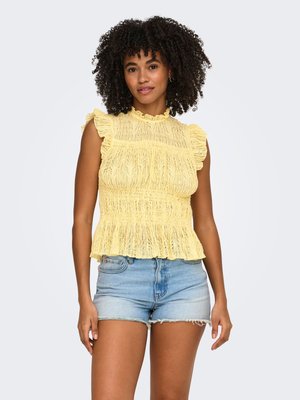 Femme aux cheveux bouclés portant un top sans manches jaune en dentelle à volants et un short en jean bleu clair sur un fond uni.