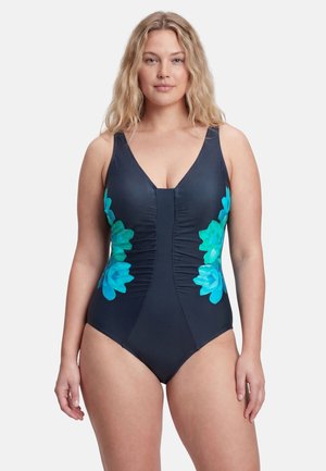 Femme blonde portant un maillot de bain une pièce noir avec des motifs floraux bleus et verts sur les côtés, debout devant un fond uni.
