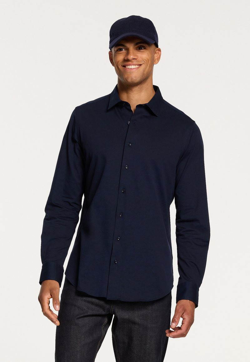 Camicia a maniche lunghe blu navy con colletto classico, texture liscia e bottoni frontali. Indossata con un cappellino blu coordinato.