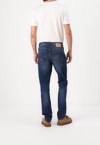 Man i blå jeans, vit kortärmad skjorta och bruna skor som står vänd bort mot en enfärgad vit bakgrund.