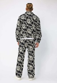 Homme avec des dreadlocks châtain clair portant une veste et un pantalon à motifs floraux noirs et blancs, se tenant devant un fond blanc uni.