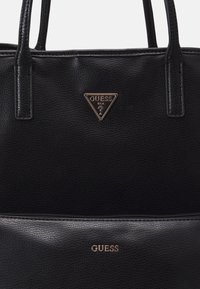 Sort lædertaske med tekstureret overflade, fremtrædende trekantet logo i guld og en lynlåslomme med det prægede mærkenavn.