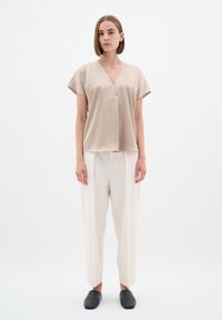 Beige satin-paita, jossa on lyhyet hihat ja V-pääntie, yhdistettynä löysiin vaalean kremppahousuihin ja mustiin liukumallisiin kenkiin.
