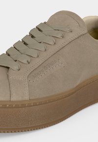 Κοντινή εικόνα ενός μπεζ σουέντ sneaker με χοντρή σόλα, ομοιόχρωμα κορδόνια και διακριτικό ανάγλυφο λογότυπο στην πλευρική επιφάνεια.