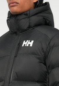 Gros plan sur une veste en duvet noire avec capuche, poche zippée sur la poitrine et logo HH blanc sur le côté gauche de la poitrine portée par une personne.