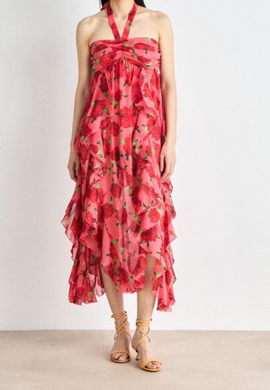 Femme portant une robe rose sans manches à col halter avec imprimé floral rouge et couches à volants, associée à des sandales à talons orange à brides.