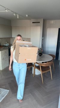 Braune Kartonbox mit Zalando-Logo, von einer Person in hellblauen Jeans gehalten, in einem modernen Innenraum mit Holzmöbeln.