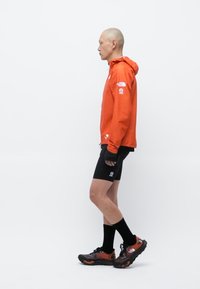Veste à capuche rouge avec des accents noirs, short noir court et chaussettes noires. Les chaussures sont sombres avec des éléments rouges et gris. Vue de côté.