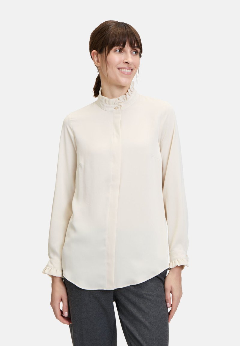 Crème blouse met een hoge ruchesboord, lange mouwen, knoopsluiting en een subtiele glans; gecombineerd met grijze broek.