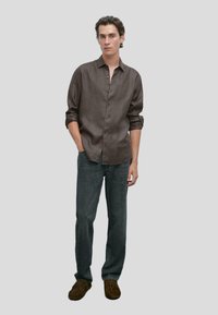 Camicia button-up in lino marrone scuro, maniche lunghe arrotolate, abbinata a jeans grigio scuro e scarpe in suede marrone. Look semplice e casual.