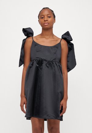 Robe en satin noire avec de grands nœuds aux bretelles, une coupe ample et une taille froncée, présentant une texture lisse et brillante.