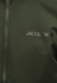 Tessuto verde oliva con una cerniera nera sul lato sinistro e il logo testuale "JACK & JONES" stampato sul lato destro.