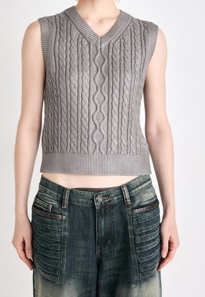 Veste sans manches en tricot gris avec motif torsadé portée avec un jean bleu délavé comportant de grandes poches avant matelassées et une bordure noire.