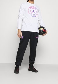 Vit långärmad skjorta med rosa Paris-logga; svarta joggers med rosa och blå detaljer; svarta sneakers med röda inslag; håller en fotboll.