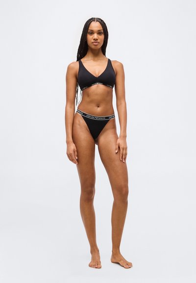 Femme debout, pieds nus, portant un ensemble de sous-vêtements noirs Emporio Armani avec ceinture à logo, sur un fond clair uni.