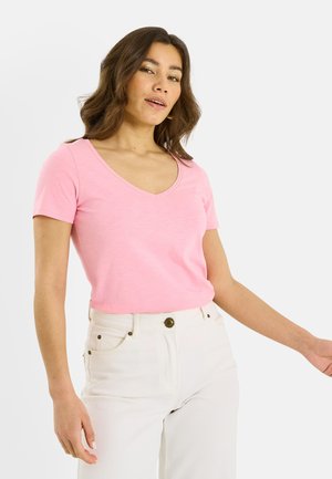 Femme portant un haut rose à col en V et manches courtes ainsi qu'un pantalon blanc taille haute, sur un fond clair uni.