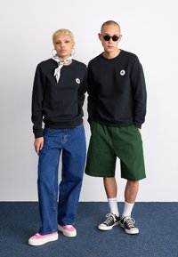 Sudaderas negras con cuello redondo, jeans holgados azules, pantalones cortos verdes, zapatillas rosas y zapatillas negras. Los accesorios incluyen una bandana con estampado.