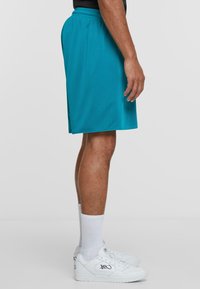 Turkise treningsshorts med mesh tekstur, elastisk linning og design som går til kneet, båret med hvite joggesko og sokker.