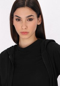 Crna zip jakna s mekanom teksturom, s kapuljačom i otvorenim dijelom preko obične crne majice. Jednostavan, minimalistički dizajn.