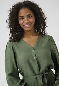 Blouse verte texturée avec un décolleté en V, un devant à boutons et des manches longues. Elle possède une taille à nouer et des détails de manches froncées.