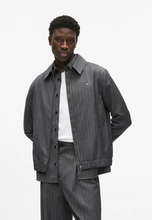 Hombre con chaqueta de rayas grises y pantalones a juego sobre camisa blanca, de pie con las manos detrás de la espalda, expresión neutral.