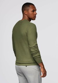 Olijfgroen gebreid sweater met lange mouwen en ribbelstructuur, voorzien van een ronde halslijn en een strakke pasvorm, gecombineerd met lichtgrijze broeken.