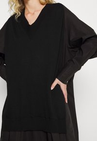 Schwarzer Strickpullover mit V-Ausschnitt und geripptem Saum, aus lockerem, strukturiertem Stoff und mit langen Ärmeln, die Knopfakzente an den Bündchen haben.