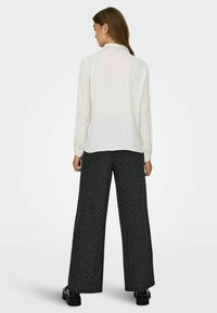 Blouse blanche à manches longues avec une texture lisse, associée à un pantalon noir large présentant un léger motif de rayures. Encolure simple avec boutons.