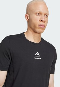Svart kortermet t-skjorte laget av myk stoff, med en hvit Adidas-logo og "LesMills"-tekst på brystet, rund hals.