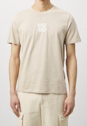 Man draagt een beige T-shirt met korte mouwen met het witte "HUCO"-logo op de borst en bijpassende beige cargobroek, staand tegen een lichte achtergrond.