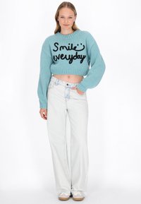 Junge Frau in einem hellblauen, kurz geschnittenen Pullover mit dem Text "Lächele jeden Tag", weißen weit geschnittenen Jeans und weißen Turnschuhen, die mit einer Hand in der Tasche steht.